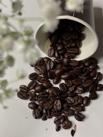 Dark roasted coffee beans are scattered across a light surface, spilling out from a tipped paper cup. Out-of-focus white and green floral elements are present in the foreground, adding a soft touch to the scene.