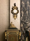 An ornate antique clock with intricate gold detailing is mounted on a richly textured wall. Below the clock is a decorative cabinet adorned with golden accents and a small statue of a seated figure. Luxurious, patterned curtains drape along the side, adding an element of sophistication to the setting.