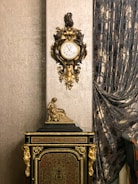 An ornate antique clock with intricate gold detailing is mounted on a richly textured wall. Below the clock is a decorative cabinet adorned with golden accents and a small statue of a seated figure. Luxurious, patterned curtains drape along the side, adding an element of sophistication to the setting.
