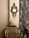 An ornate antique clock with intricate gold detailing is mounted on a richly textured wall. Below the clock is a decorative cabinet adorned with golden accents and a small statue of a seated figure. Luxurious, patterned curtains drape along the side, adding an element of sophistication to the setting.
