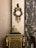 An ornate antique clock with intricate gold detailing is mounted on a richly textured wall. Below the clock is a decorative cabinet adorned with golden accents and a small statue of a seated figure. Luxurious, patterned curtains drape along the side, adding an element of sophistication to the setting.