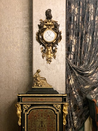 An ornate antique clock with intricate gold detailing is mounted on a richly textured wall. Below the clock is a decorative cabinet adorned with golden accents and a small statue of a seated figure. Luxurious, patterned curtains drape along the side, adding an element of sophistication to the setting.