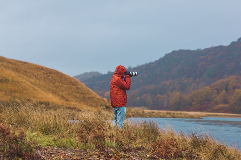 man in red jacket standing on grass field near body of water during daytime