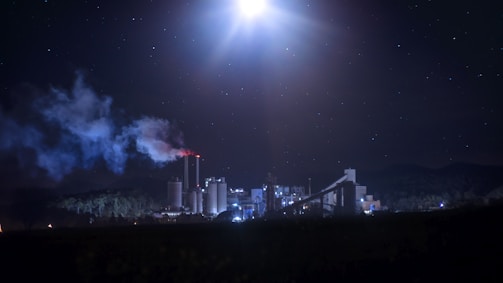 A nighttime industrial scene with a factory emitting smoke from tall chimneys. The factory is illuminated against a dark sky filled with stars. The moon or a bright light source is visible, casting light over the landscape. The surrounding area is largely in shadow, with faint outlines of trees and possibly mountains in the background.