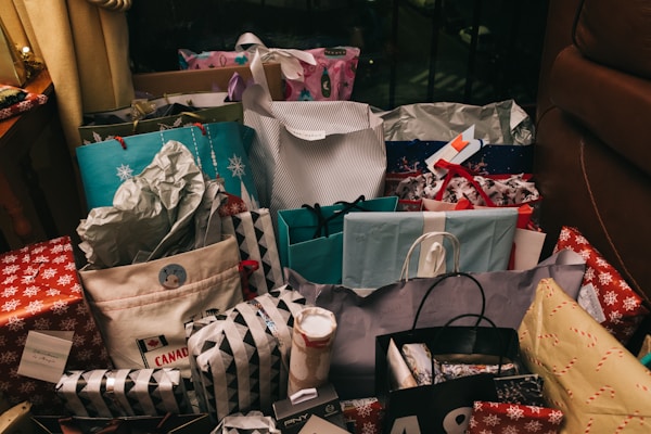 A collection of various wrapped gifts and shopping bags is placed on the floor. The presents are wrapped in colorful paper and decorated with ribbons. Some bags display festive designs, and nearby are decorative items enhancing the holiday theme.