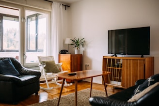 A cozy living room with a comfortable leather sofa, a wooden rocking chair, and a large flat-screen television on a wooden cabinet. There is a small coffee table in the center, a potted plant, and a lamp on a side table. Natural light streams in from the large window, illuminating the room.