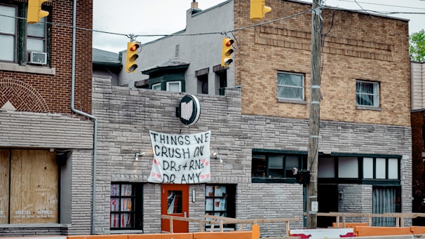 A street scene depicting a brick and stone building with a sign that reads 'Things We Crush On Drs & RNs Dr Amy'. The building has boarded-up windows and a small circular logo above the sign. There are overhead power lines and a traffic light with red and yellow signals. An orange barricade is partially visible at the bottom of the image.