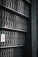 A series of metal gates with a mesh and diamond pattern arranged vertically, casting shadows on the wall behind. There's a no parking sign attached to one side of the gate.