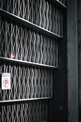 A series of metal gates with a mesh and diamond pattern arranged vertically, casting shadows on the wall behind. There's a no parking sign attached to one side of the gate.