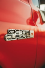 Close-up view of a red vehicle featuring a metallic emblem with the text 'Sierra Classic'. The surface of the car appears smooth and reflective, highlighting the shiny chrome detail of the badge.