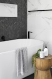 A modern bathroom features a pristine white bathtub with a light gray patterned towel draped over the edge. Adjacent to the tub is a wooden stool holding a candle, a plant, and a soap dispenser. The wall features a contrast of white marble tiles and dark herringbone tiles.