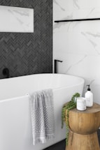 A modern bathroom features a pristine white bathtub with a light gray patterned towel draped over the edge. Adjacent to the tub is a wooden stool holding a candle, a plant, and a soap dispenser. The wall features a contrast of white marble tiles and dark herringbone tiles.