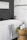 A modern bathroom features a pristine white bathtub with a light gray patterned towel draped over the edge. Adjacent to the tub is a wooden stool holding a candle, a plant, and a soap dispenser. The wall features a contrast of white marble tiles and dark herringbone tiles.