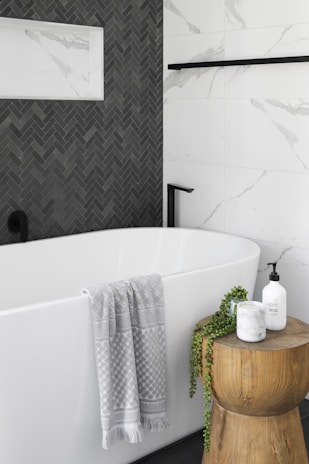 A modern bathroom features a pristine white bathtub with a light gray patterned towel draped over the edge. Adjacent to the tub is a wooden stool holding a candle, a plant, and a soap dispenser. The wall features a contrast of white marble tiles and dark herringbone tiles.