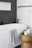 A modern bathroom features a pristine white bathtub with a light gray patterned towel draped over the edge. Adjacent to the tub is a wooden stool holding a candle, a plant, and a soap dispenser. The wall features a contrast of white marble tiles and dark herringbone tiles.
