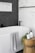 A modern bathroom features a pristine white bathtub with a light gray patterned towel draped over the edge. Adjacent to the tub is a wooden stool holding a candle, a plant, and a soap dispenser. The wall features a contrast of white marble tiles and dark herringbone tiles.