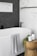 A modern bathroom features a pristine white bathtub with a light gray patterned towel draped over the edge. Adjacent to the tub is a wooden stool holding a candle, a plant, and a soap dispenser. The wall features a contrast of white marble tiles and dark herringbone tiles.
