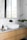 A modern bathroom countertop features a rectangular white vessel sink with a black faucet above it. Adjacent to the sink, a wooden tray displays a black bottle, a scrub brush, and a folded cloth. On the right side, a glass vase holds green monstera leaves. The countertop itself is sleek and grey, set against a backdrop of marble tiles and a large mirror, creating a clean and minimalist aesthetic.