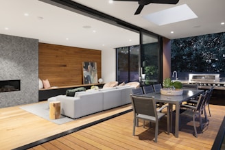 A modern living space seamlessly blends indoor and outdoor elements, featuring a cozy lounge area with a sectional sofa and pillows, a contemporary dining table set, and a built-in outdoor grill. The space is accented with a wooden wall, abstract art, and plants. A large sliding glass door and a skylight allow ample natural light.
