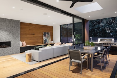 A modern living space seamlessly blends indoor and outdoor elements, featuring a cozy lounge area with a sectional sofa and pillows, a contemporary dining table set, and a built-in outdoor grill. The space is accented with a wooden wall, abstract art, and plants. A large sliding glass door and a skylight allow ample natural light.