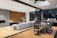 A modern living space seamlessly blends indoor and outdoor elements, featuring a cozy lounge area with a sectional sofa and pillows, a contemporary dining table set, and a built-in outdoor grill. The space is accented with a wooden wall, abstract art, and plants. A large sliding glass door and a skylight allow ample natural light.