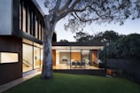 A modern house with a sleek architectural design featuring large glass windows and wooden accents. The outdoor area includes a deck with a dining table and chairs next to a barbecue grill, framed by a large tree and lush greenery.
