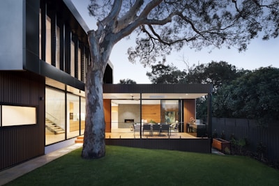A modern house with a sleek architectural design featuring large glass windows and wooden accents. The outdoor area includes a deck with a dining table and chairs next to a barbecue grill, framed by a large tree and lush greenery.