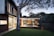 A modern house with a sleek architectural design featuring large glass windows and wooden accents. The outdoor area includes a deck with a dining table and chairs next to a barbecue grill, framed by a large tree and lush greenery.