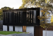 A modern building with a sleek design featuring dark, reflective panels and large, vertical window shades. The structure is surrounded by a well-maintained lawn and a few trees, and the sun casts soft shadows, highlighting the architectural lines.