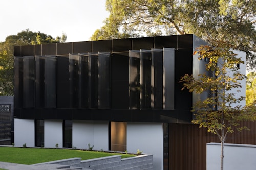 A modern building with a sleek design featuring dark, reflective panels and large, vertical window shades. The structure is surrounded by a well-maintained lawn and a few trees, and the sun casts soft shadows, highlighting the architectural lines.