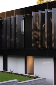 A modern architectural detail featuring a dark, sleek facade with vertical panels. Below, a minimalist garden area with a low stone wall and neatly trimmed grass is visible. A large window reveals a softly lit interior space.