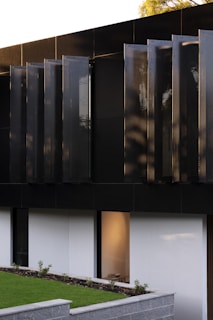 A modern architectural detail featuring a dark, sleek facade with vertical panels. Below, a minimalist garden area with a low stone wall and neatly trimmed grass is visible. A large window reveals a softly lit interior space.