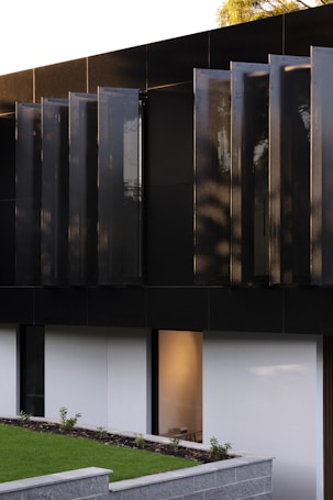 A modern architectural detail featuring a dark, sleek facade with vertical panels. Below, a minimalist garden area with a low stone wall and neatly trimmed grass is visible. A large window reveals a softly lit interior space.