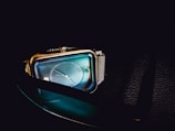 A close-up view of a stylish rectangular wristwatch with a blue iridescent face, silver metal casing, and a mesh wristband. The background is dark, enhancing the watch's sleek and modern design.