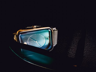 A close-up view of a stylish rectangular wristwatch with a blue iridescent face, silver metal casing, and a mesh wristband. The background is dark, enhancing the watch's sleek and modern design.