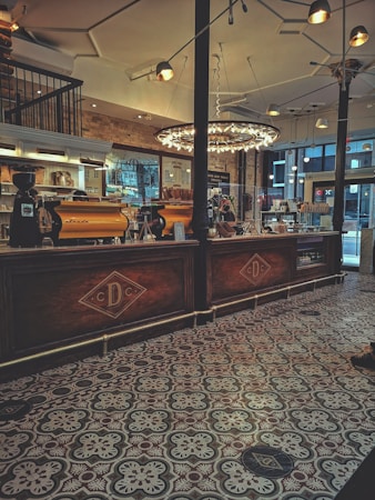 A cozy caf&eacute; interior features ornate patterned tile flooring and a warmly lit counter area with two espresso machines. The ambient lighting includes a large, circular chandelier hanging from the ceiling. Behind the counter, there are shelves displaying various items and a brick wall adorned with framed artwork and a menu. The space is designed with a mix of modern and rustic elements, creating an inviting atmosphere.