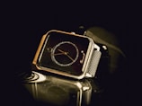 A square-faced watch with a black dial and gold hands is displayed prominently against a dark background. The metal mesh band of the watch is shiny and reflective. The surface on which the watch rests is also reflective, adding a slight mirrored effect.