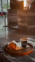 A croissant and a cup of coffee are placed on a round wooden tray at a marble counter. The setting appears to be inside a modern cafe with a sleek interior design. The lighting is warm, giving the atmosphere a cozy and inviting feel.