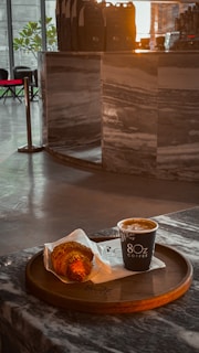 A croissant and a cup of coffee are placed on a round wooden tray at a marble counter. The setting appears to be inside a modern cafe with a sleek interior design. The lighting is warm, giving the atmosphere a cozy and inviting feel.