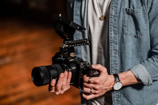 A person holding a professional camera with both hands. The camera has a large lens and a complex rig setup, indicating it might be used for filming. The individual is wearing a denim shirt and a wristwatch. The setting suggests a warm, wooden background possibly in a studio or workshop.