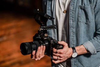 A person holding a professional camera with both hands. The camera has a large lens and a complex rig setup, indicating it might be used for filming. The individual is wearing a denim shirt and a wristwatch. The setting suggests a warm, wooden background possibly in a studio or workshop.