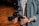 A person holding a professional camera with both hands. The camera has a large lens and a complex rig setup, indicating it might be used for filming. The individual is wearing a denim shirt and a wristwatch. The setting suggests a warm, wooden background possibly in a studio or workshop.