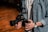 A person holding a professional camera with both hands. The camera has a large lens and a complex rig setup, indicating it might be used for filming. The individual is wearing a denim shirt and a wristwatch. The setting suggests a warm, wooden background possibly in a studio or workshop.
