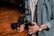 A person holding a professional camera with both hands. The camera has a large lens and a complex rig setup, indicating it might be used for filming. The individual is wearing a denim shirt and a wristwatch. The setting suggests a warm, wooden background possibly in a studio or workshop.