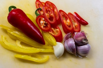 An arrangement of fresh vegetables and garlic cloves on a light-colored cutting board. On the left, bright yellow bell pepper strips are placed alongside a whole red bell pepper. Sliced red jalapeños are neatly aligned next to whole and peeled garlic cloves.
