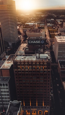An aerial view of a cityscape at dusk, featuring several tall buildings with prominent signage, including one for Chase Bank. The skyline is illuminated by the setting sun, casting warm hues over the city. Roads run between the buildings, and the horizon is visible in the distance.
