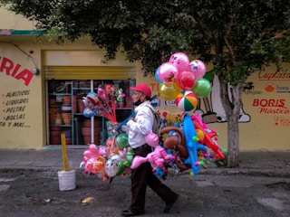 A street vendor is walking along a sidewalk carrying a large assortment of colorful balloons and inflatable toys. The scene is set against a vibrant wall with various advertisements and a few trees providing shade. The vendor is wearing a red cap and a white shirt, contributing to the colorful atmosphere of the scene.