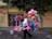 A street vendor is walking along a sidewalk carrying a large assortment of colorful balloons and inflatable toys. The scene is set against a vibrant wall with various advertisements and a few trees providing shade. The vendor is wearing a red cap and a white shirt, contributing to the colorful atmosphere of the scene.