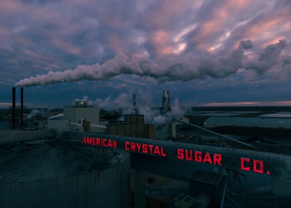 A factory complex emitting large clouds of smoke under a dramatic, cloudy sky. The scene is dominated by an industrial environment with metal structures and chimneys releasing emissions into the atmosphere. Bright red neon signage reads 'AMERICAN CRYSTAL SUGAR CO.' at the forefront.