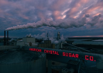 A factory complex emitting large clouds of smoke under a dramatic, cloudy sky. The scene is dominated by an industrial environment with metal structures and chimneys releasing emissions into the atmosphere. Bright red neon signage reads 'AMERICAN CRYSTAL SUGAR CO.' at the forefront.